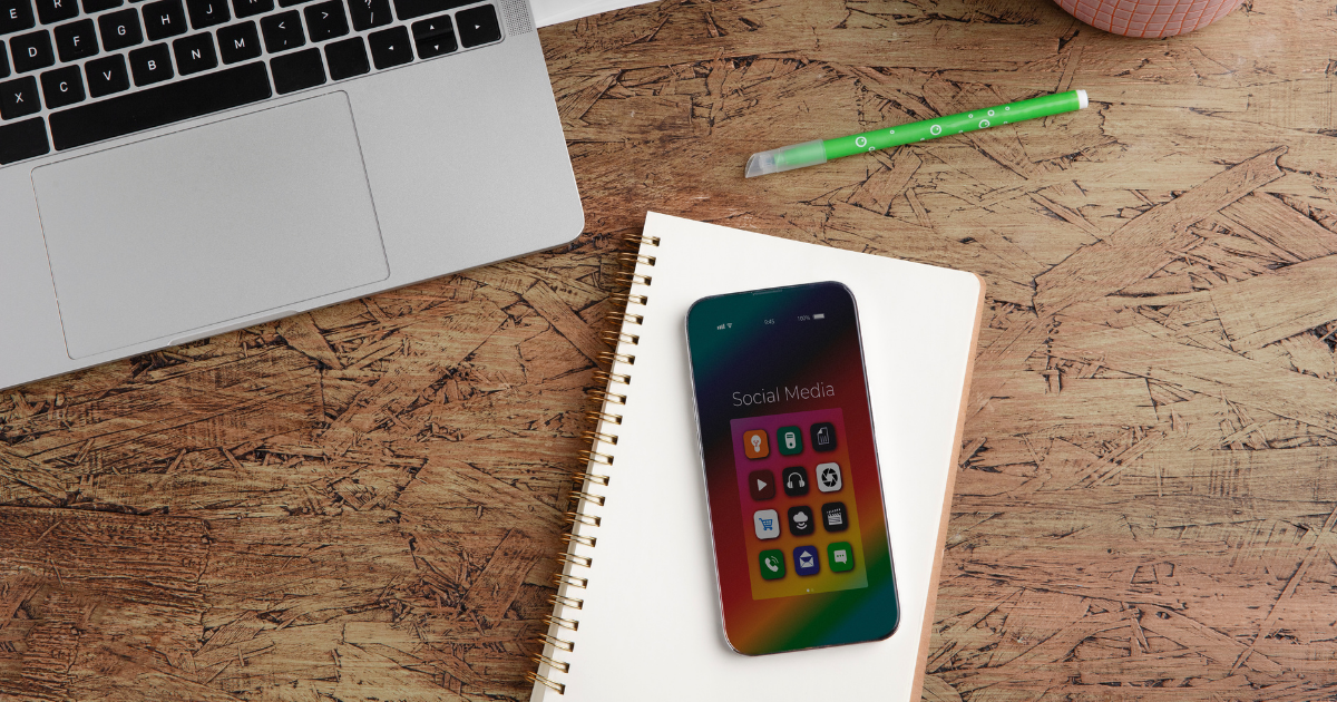 Ein Smartphone liegt auf einem Spiralnotizbuch auf einem Holztisch, neben einem offenen Laptop, einer Kaffeetasse, einem grünen Stift und einer Topfpflanze. Auf dem Display des Smartphones ist ein Bildschirm mit verschiedenen Social-Media-App-Icons zu sehen.