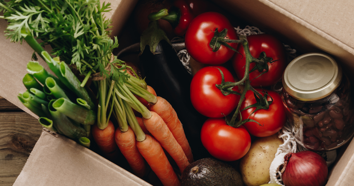 Offene Pappschachtel gefüllt mit frischem Gemüse und Obst, darunter Karotten, Tomaten, Frühlingszwiebeln, Avocado, Apfel, Kartoffel, Aubergine sowie ein Glas mit eingelegten Bohnen.