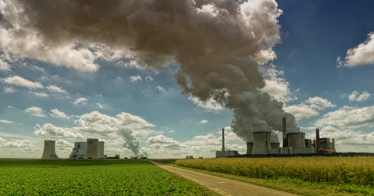 Großes Kraftwerk mit mehreren Kühltürmen, aus denen dichte Rauch- und Dampfwolken aufsteigen; davor Felder und ein Weg unter blauem Himmel mit Wolken.