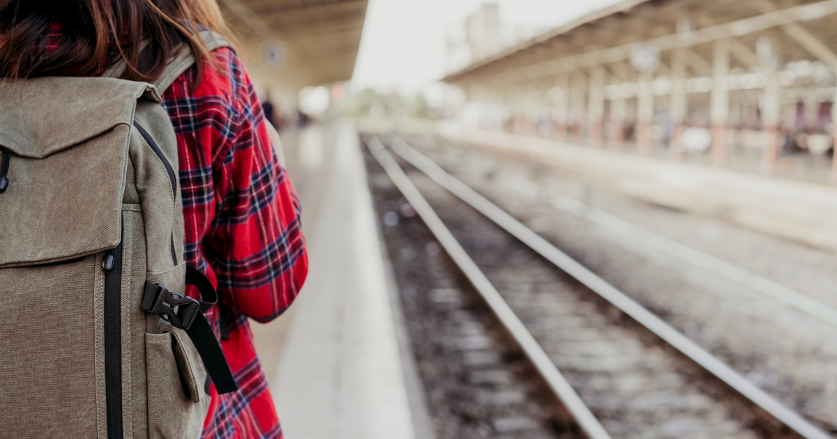Person mit Rucksack und rotem Flanellhemd steht auf einem Bahnsteig neben den Gleisen.