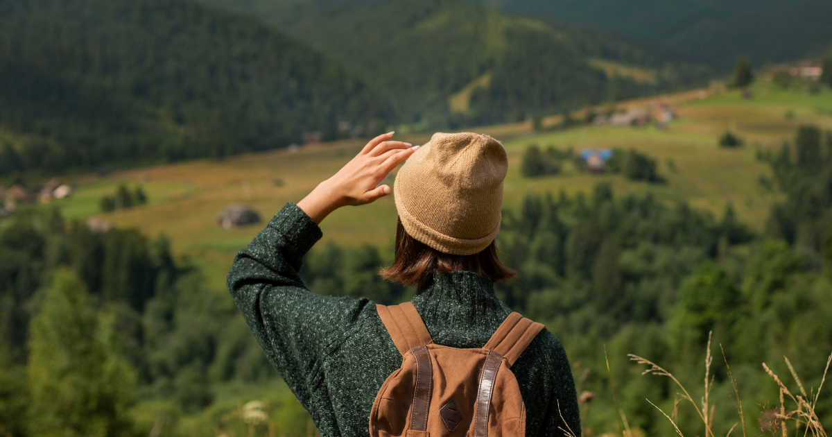 Eine Person mit Rucksack und Mütze steht auf einem Hügel und blickt in eine weite, grüne Berglandschaft.