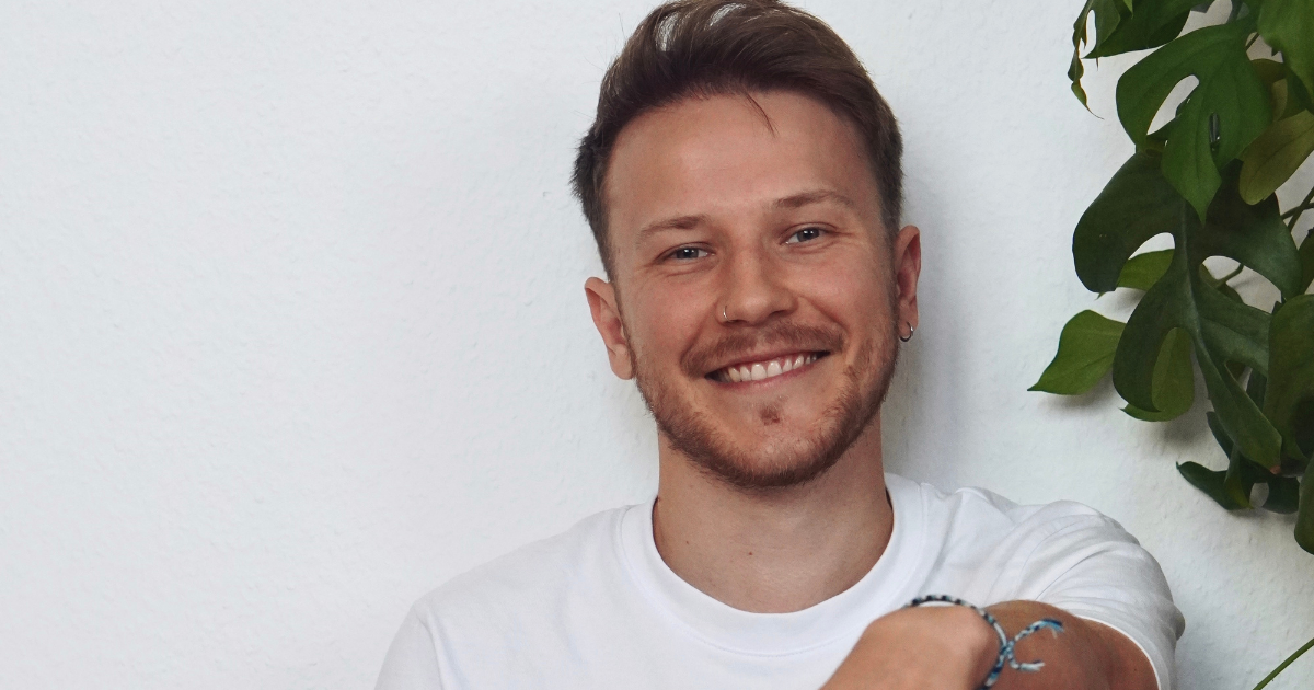 Influencer henreymisterio sitzt lächelnd in weißem T-Shirt vor einer weißen Wand neben einer Zimmerpflanze und blickt in die Kamera.