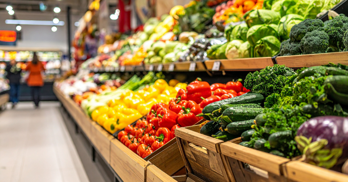 Frische Gemüseauslage in einem Supermarkt mit einer Vielzahl an bunten Paprika, Zucchini, Brokkoli, Kohl und weiteren Gemüsesorten in Holzkisten und Regalen.