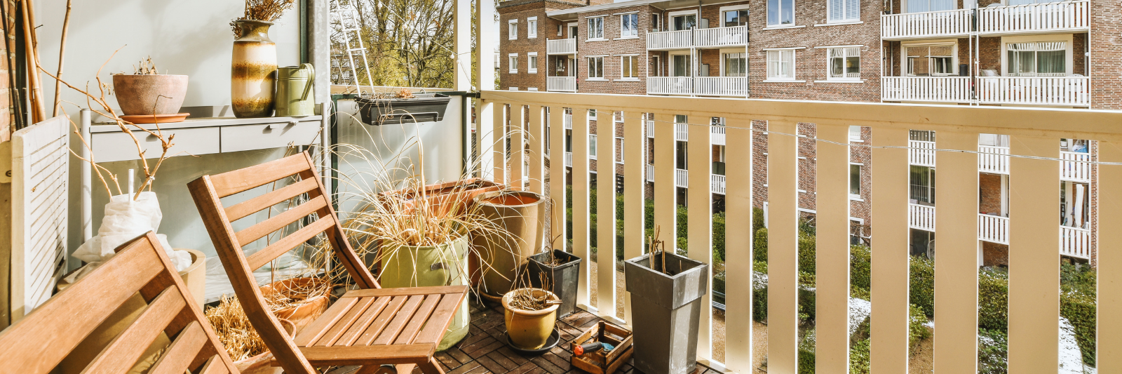Kleiner, sonniger Balkon mit Holzboden, zwei Holzstühlen, verschiedenen Pflanzgefäßen und dekorativen Vasen; im Hintergrund sind die Balkone eines mehrstöckigen Wohnhauses zu sehen.