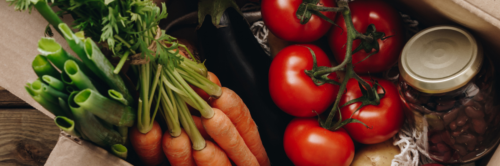 Offene Pappschachtel gefüllt mit frischem Gemüse und Obst, darunter Karotten, Tomaten, Frühlingszwiebeln, Avocado, Apfel, Kartoffel, Aubergine sowie ein Glas mit eingelegten Bohnen.