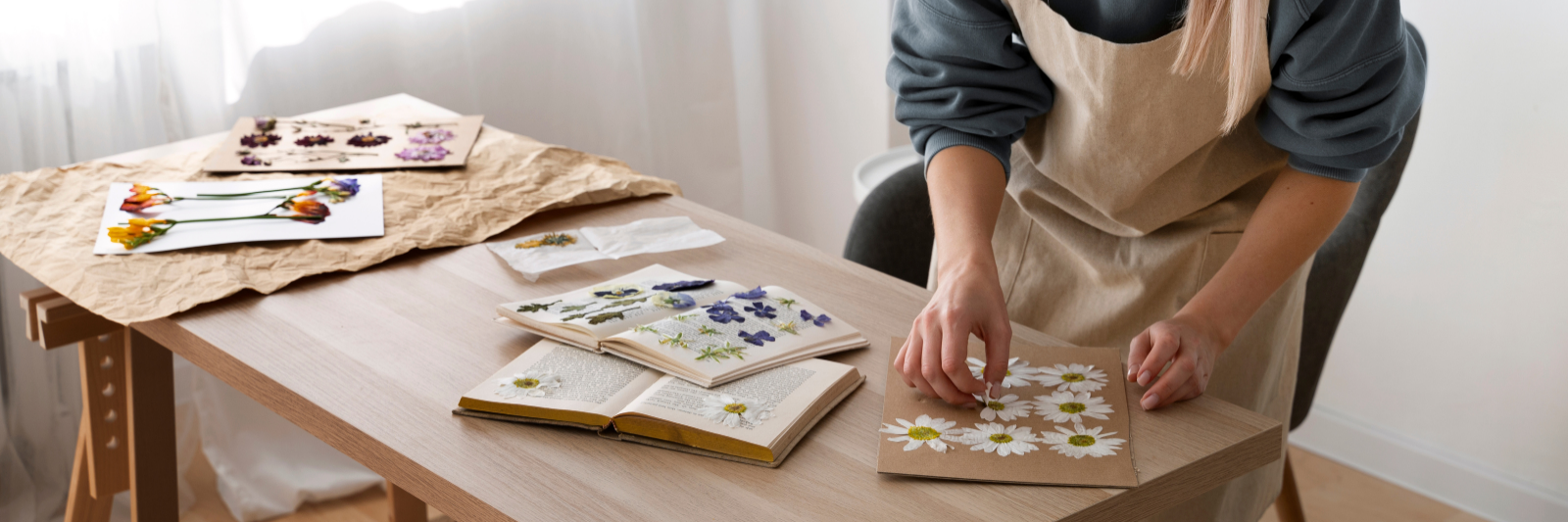 Eine Frau in Schürze arbeitet an einem Tisch und arrangiert gepresste Blumen auf einer Pappe. Auf dem Tisch liegen weitere gepresste Blüten und geöffnete Bücher mit Pflanzenmustern.