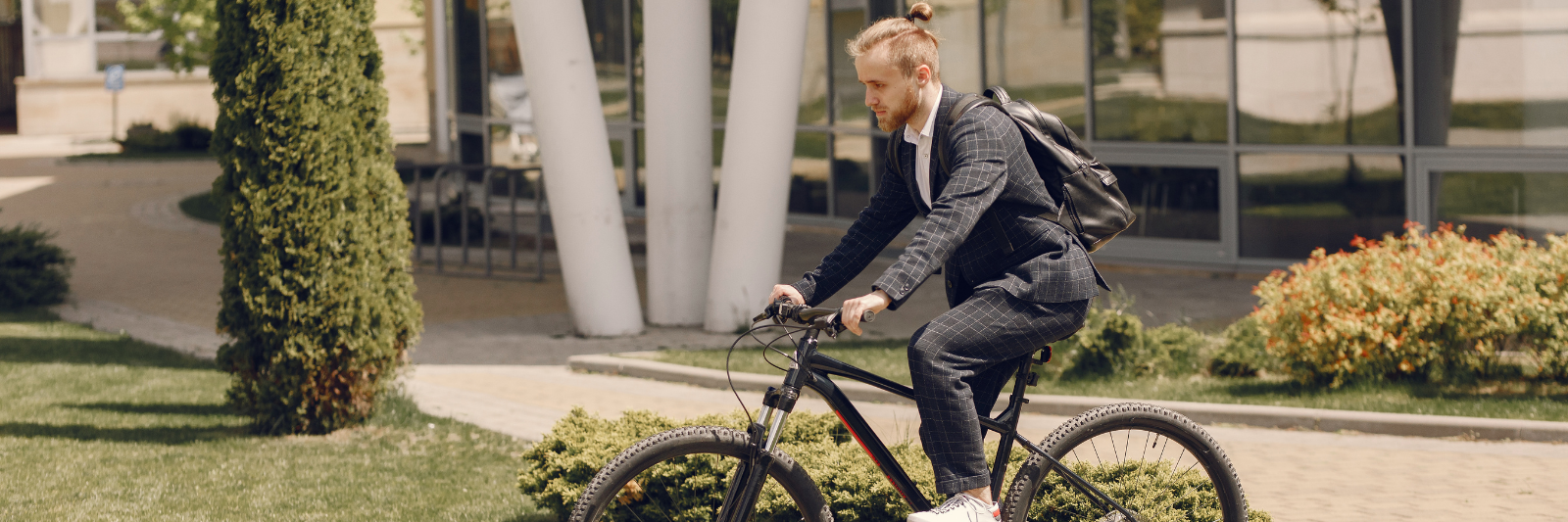 Ein Mann in Anzug und Rucksack fährt auf einem schwarzen Fahrrad durch eine moderne, begrünte Außenanlage vor einem Bürogebäude.
