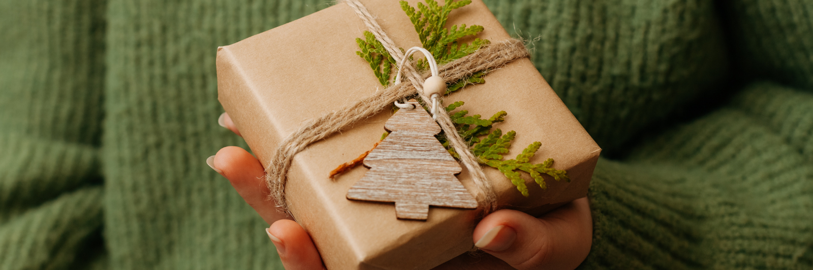 Hände halten ein naturbelassen verpacktes Geschenk mit braunem Papier, Juteschnur, grünem Zweig und einem kleinen Holzanhänger in Tannenbaumform.