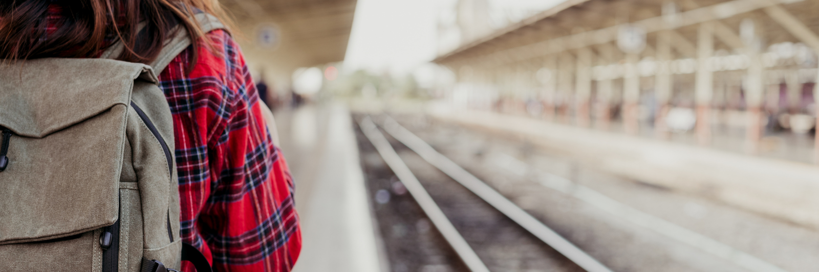 Person mit Rucksack und rotem Flanellhemd steht auf einem Bahnsteig neben den Gleisen.