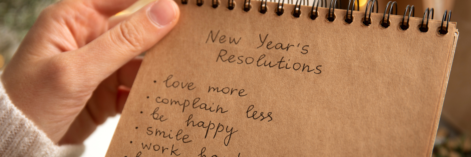 Hand hält ein Notizbuch mit der Überschrift „New Year's Resolutions“ und einer Liste guter Vorsätze, umgeben von warmen Lichtern im Hintergrund.