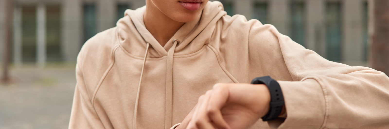 Eine junge Person mit beigefarbenem Hoodie schaut im Freien auf ihre Smartwatch.