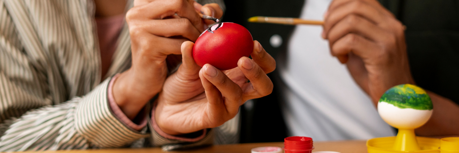 Zwei Personen bemalen gemeinsam ein rotes Osterei; eine Hand hält das Ei, während die andere feine Details mit einem Pinsel aufträgt. Im Vordergrund stehen offene Farbtöpfe und ein weiteres bemaltes Ei auf einem Ständer.
