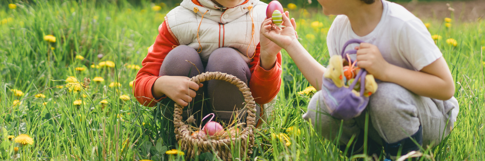 Zwei Kinder mit Osterkostümen suchen im grünen Gras nach Ostereiern. Ein Kind trägt eine gelbe Kükenmaske, das andere Hasenohren, und sie halten bunte Eier in den Händen.