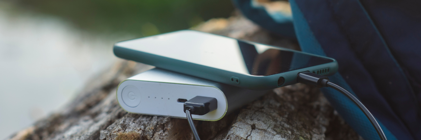 Ein Smartphone liegt auf einer Powerbank, die über ein Kabel angeschlossen ist. Beides befindet sich auf einem Baumstumpf im Freien, neben einem Rucksack.