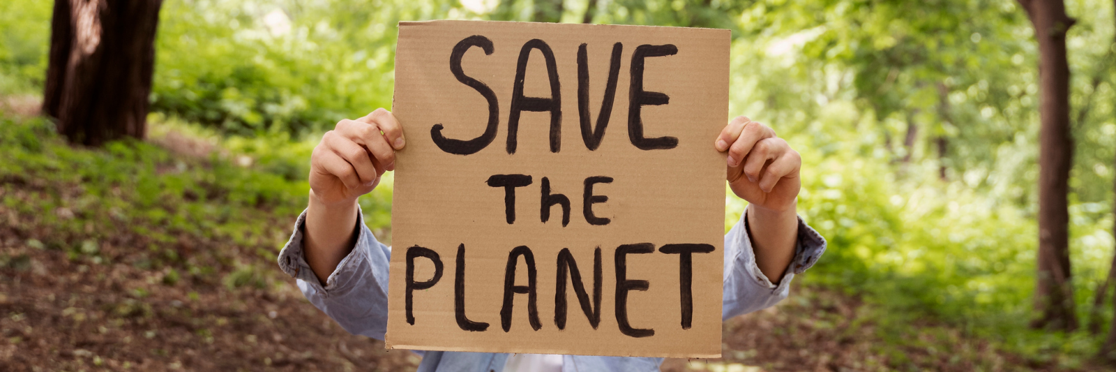 Eine sitzende Person hält ein Pappschild mit der Aufschrift „SAVE THE PLANET“ vor ihrem Gesicht, umgeben von Bäumen in einem Wald; neben ihr liegt ein weiteres Schild mit Umweltbotschaft.