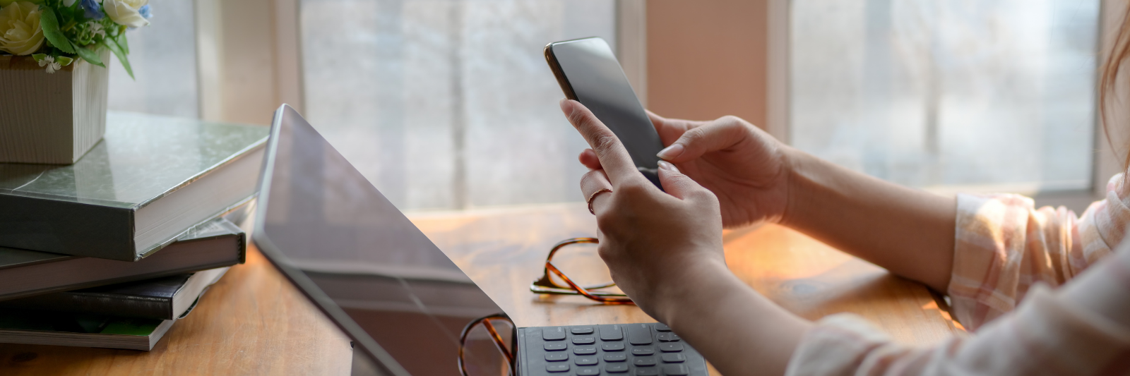 Person sitzt am Schreibtisch vor einem aufgeklappten Laptop und hält ein Smartphone in den Händen; auf dem Tisch liegen Bücher, eine Brille und eine Tasse Kaffee, im Hintergrund große Fenster mit Tageslicht.