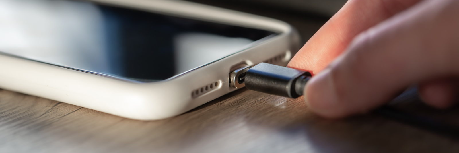 Nahaufnahme einer Hand, die ein Ladekabel in den Anschluss eines Smartphones mit weißer Hülle steckt, liegend auf einer Holzoberfläche.