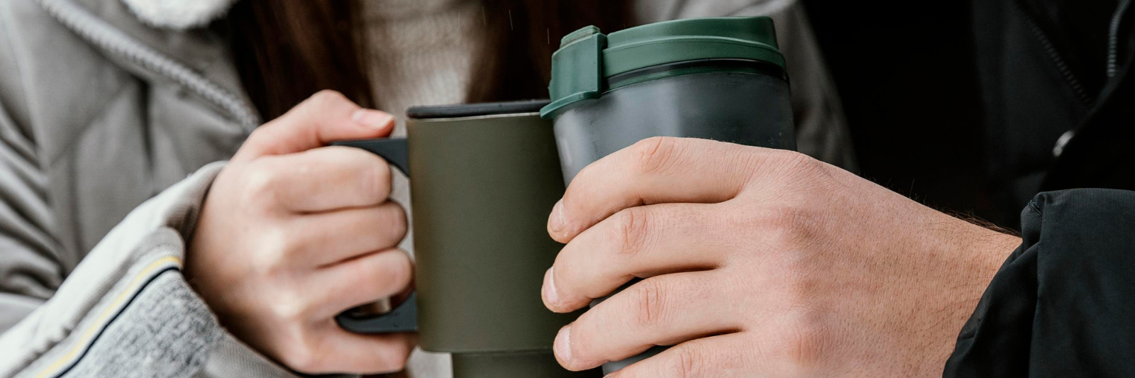 Zwei Personen stoßen im Freien mit Thermobechern und einer Tasse an. Beide tragen warme Kleidung, und der Fokus liegt auf den Händen und den Trinkgefäßen.