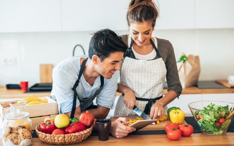 Ein lächelndes Paar in der Küche, beide mit Schürzen, bereitet gemeinsam eine Mahlzeit zu; die Frau schneidet gelbe Paprika auf einem Schneidebrett, während der Mann danebensteht und zuschaut, umgeben von frischem Obst und Gemüse.