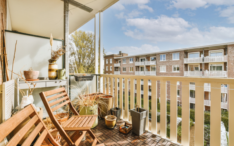 Kleiner, sonniger Balkon mit Holzboden, zwei Holzstühlen, verschiedenen Pflanzgefäßen und dekorativen Vasen; im Hintergrund sind die Balkone eines mehrstöckigen Wohnhauses zu sehen.