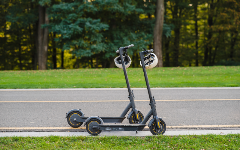 Zwei geparkte E-Scooter mit angehängten Fahrradhelmen stehen am Straßenrand vor einer Grünfläche mit Bäumen im Hintergrund.