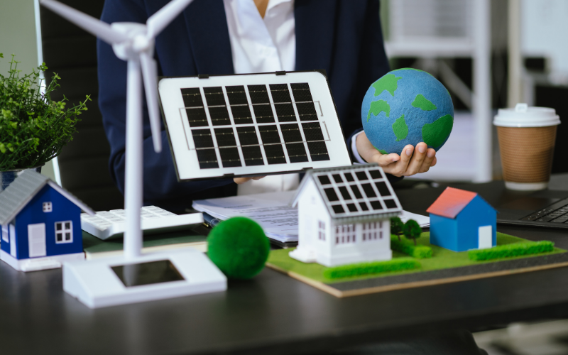 Person in Businesskleidung sitzt an einem Schreibtisch und hält ein Modell eines Solarpanels sowie eine kleine Erdkugel. Auf dem Tisch stehen Miniaturhäuser mit Solardächern, ein Modell-Windrad, Pflanzen und weitere Symbole für erneuerbare Energien.