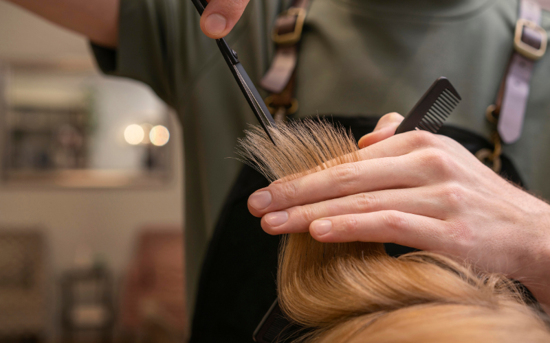 Friseur schneidet einem Kunden oder einer Kundin blonde Haarspitzen mit einer Schere, während er die Haare zwischen den Fingern hält; ein Kamm liegt in der Hand bereit.