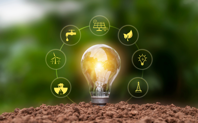 Eine leuchtende Glühbirne steht auf Erde, umgeben von kreisförmigen Symbolen zu erneuerbaren Energien wie Solarenergie, Windkraft, Wassernutzung und nachhaltigen Ressourcen, vor einem grünen Naturhintergrund.