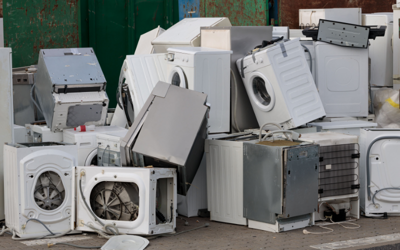 Haufen ausgedienter Haushaltsgeräte wie Waschmaschinen, Trockner und Geschirrspüler auf einem Recycling- oder Entsorgungsplatz.