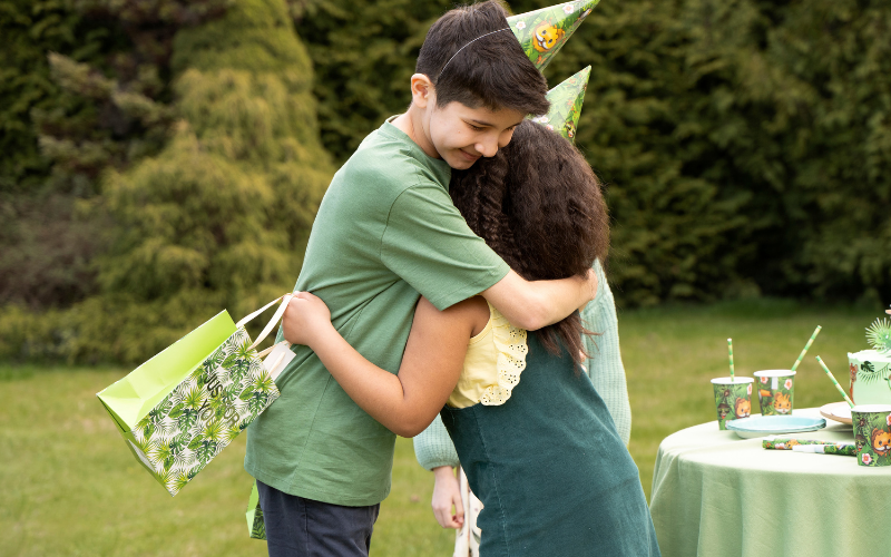 Zwei Kinder mit Partyhütchen umarmen sich im Garten; eines hält eine Geschenktüte, im Hintergrund steht ein gedeckter Geburtstagstisch mit grüner Deko.