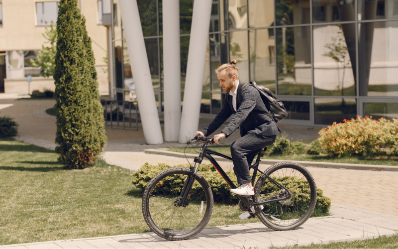 Ein Mann in Anzug und Rucksack fährt auf einem schwarzen Fahrrad durch eine moderne, begrünte Außenanlage vor einem Bürogebäude.