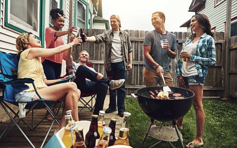 Eine Gruppe junger Erwachsener sitzt und steht fröhlich im Garten, während sie gemeinsam grillen. Einige stoßen mit Getränken an, andere unterhalten sich, und auf dem Grill liegen verschiedene Speisen. Die Szene wirkt entspannt und sommerlich.