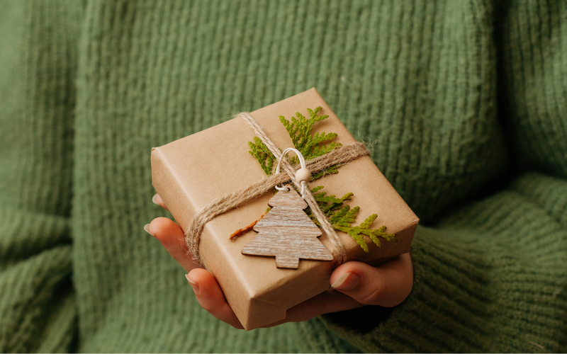 Hände halten ein naturbelassen verpacktes Geschenk mit braunem Papier, Juteschnur, grünem Zweig und einem kleinen Holzanhänger in Tannenbaumform.