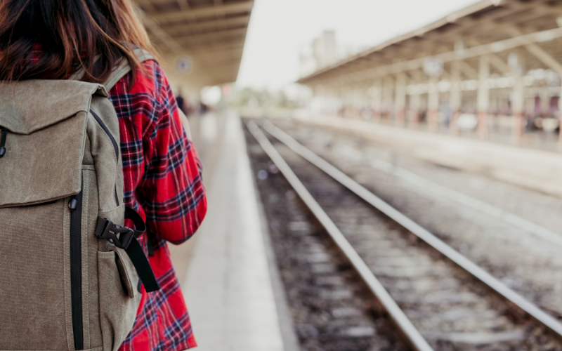 Person mit Rucksack und rotem Flanellhemd steht auf einem Bahnsteig neben den Gleisen.