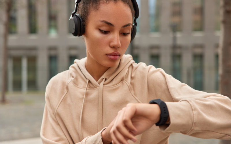 Eine junge Person mit Kopfhörern und beigefarbenem Hoodie schaut im Freien auf ihre Smartwatch.