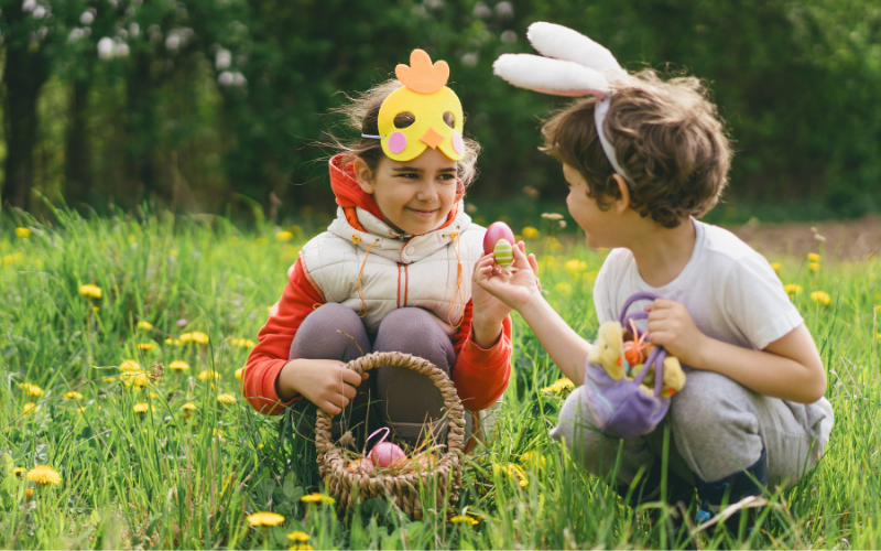 Zwei Kinder mit Osterkostümen suchen im grünen Gras nach Ostereiern. Ein Kind trägt eine gelbe Kükenmaske, das andere Hasenohren, und sie halten bunte Eier in den Händen.