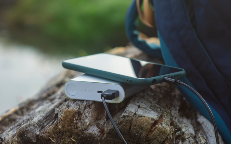 Ein Smartphone liegt auf einer Powerbank, die über ein Kabel angeschlossen ist. Beides befindet sich auf einem Baumstumpf im Freien, neben einem Rucksack.