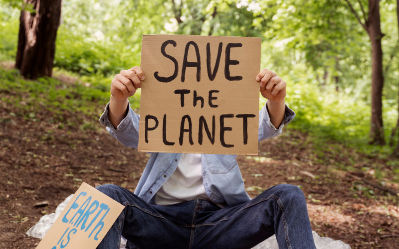 Eine sitzende Person hält ein Pappschild mit der Aufschrift „SAVE THE PLANET“ vor ihrem Gesicht, umgeben von Bäumen in einem Wald; neben ihr liegt ein weiteres Schild mit Umweltbotschaft.
