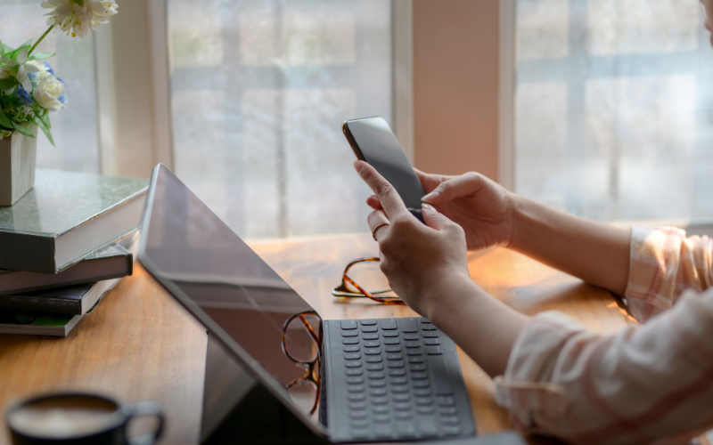 Person sitzt am Schreibtisch vor einem aufgeklappten Laptop und hält ein Smartphone in den Händen; auf dem Tisch liegen Bücher, eine Brille und eine Tasse Kaffee, im Hintergrund große Fenster mit Tageslicht.