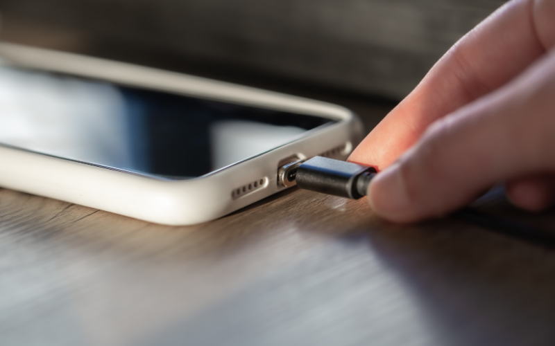 Nahaufnahme einer Hand, die ein Ladekabel in den Anschluss eines Smartphones mit weißer Hülle steckt, liegend auf einer Holzoberfläche.
