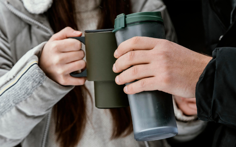 Zwei Personen stoßen im Freien mit Thermobechern und einer Tasse an. Beide tragen warme Kleidung, und der Fokus liegt auf den Händen und den Trinkgefäßen.