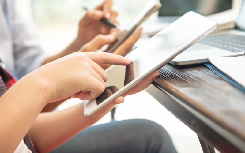 Zwei Personen sitzen an einem Tisch und arbeiten mit digitalen Geräten; im Vordergrund tippt jemand auf ein Tablet, während im Hintergrund eine weitere Hand ein Smartphone hält.