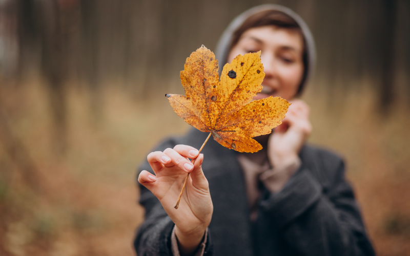 Eine Person hält ein großes, orange-gelbes Herbstblatt in den Vordergrund der Kamera. Der Hintergrund ist unscharf, zeigt aber einen Wald mit herbstlichen Farben. Die Person trägt eine graue Jacke und eine Mütze und lächelt leicht.