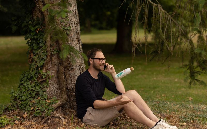 Mann sitzt am Baum auf einer Wiese und telefoniert