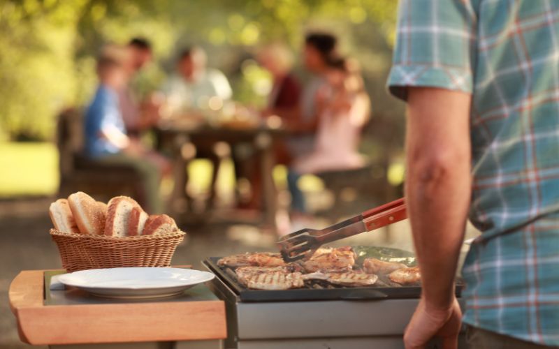 Mann grillt Fleisch, im Hintergrund Familie