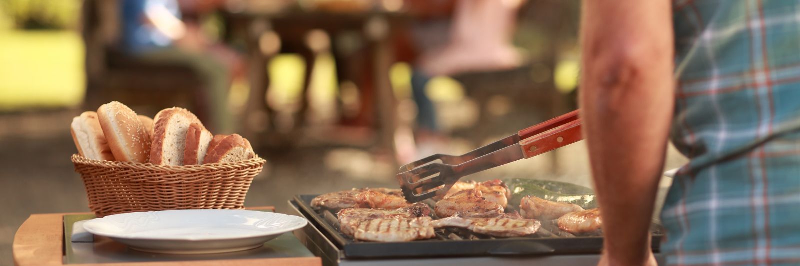 Mann grillt Fleisch, im Hintergrund Familie