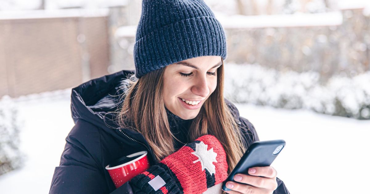 Eine Frau schaut auf ihr Handy, hinter ihr ein verschneiter Garten mit Mauer