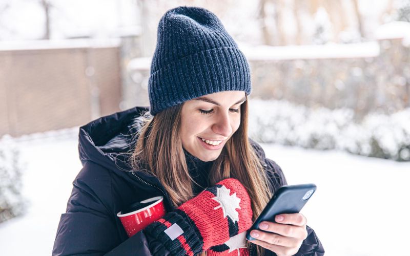 Eine Frau schaut auf ihr Handy, hinter ihr ein verschneiter Garten mit Mauer