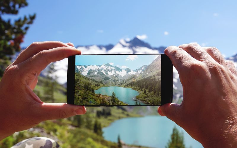 Mensch macht Foto mit Smartphone, im Hintergrund Berge, Wälder und See