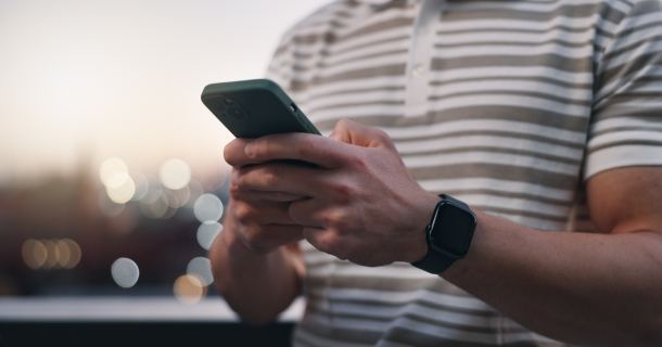 Nahaufnahme einer Person im gestreiften T-Shirt, die ein Smartphone mit beiden Händen bedient und eine schwarze Smartwatch trägt. Im Hintergrund sind unscharfe Lichter zu sehen.